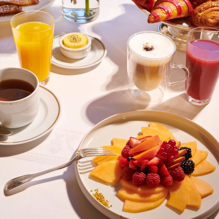 Hôtel Plaza Athénée, Paris的室内早餐。餐桌上用叉子挑水果，水果盘上放着浆果橙汁和卡布奇诺。