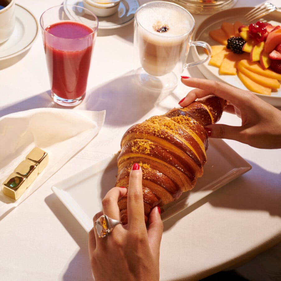 Hôtel Plaza Athénée, Paris的室内早餐。桌上用一只手挑着一个大羊角包，水果盘，浆果汁和卡布奇诺放在一边。