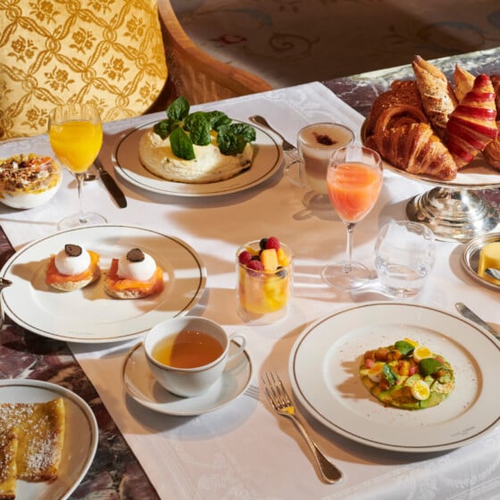 我们的主餐厅Hôtel Plaza Athénée的早餐桌，放在白色桌布上，有牛油果、白色煎蛋卷、松露鸡蛋、可丽饼和糕点，旁边还有饮料。