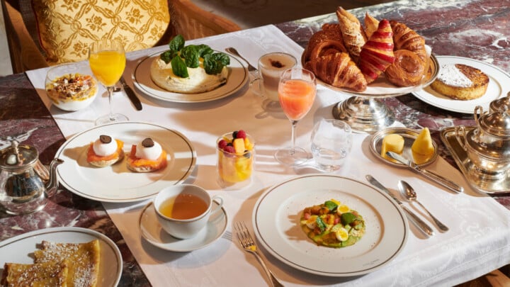 Petit-déjeuner en chambre à l'Hôtel Plaza Athénée，巴黎。Table avec une fourchette piochant des fruits dans une assiette de fruits, crêpe, ufs, juus de fruits et d'orange et cappuccino sur le côté。